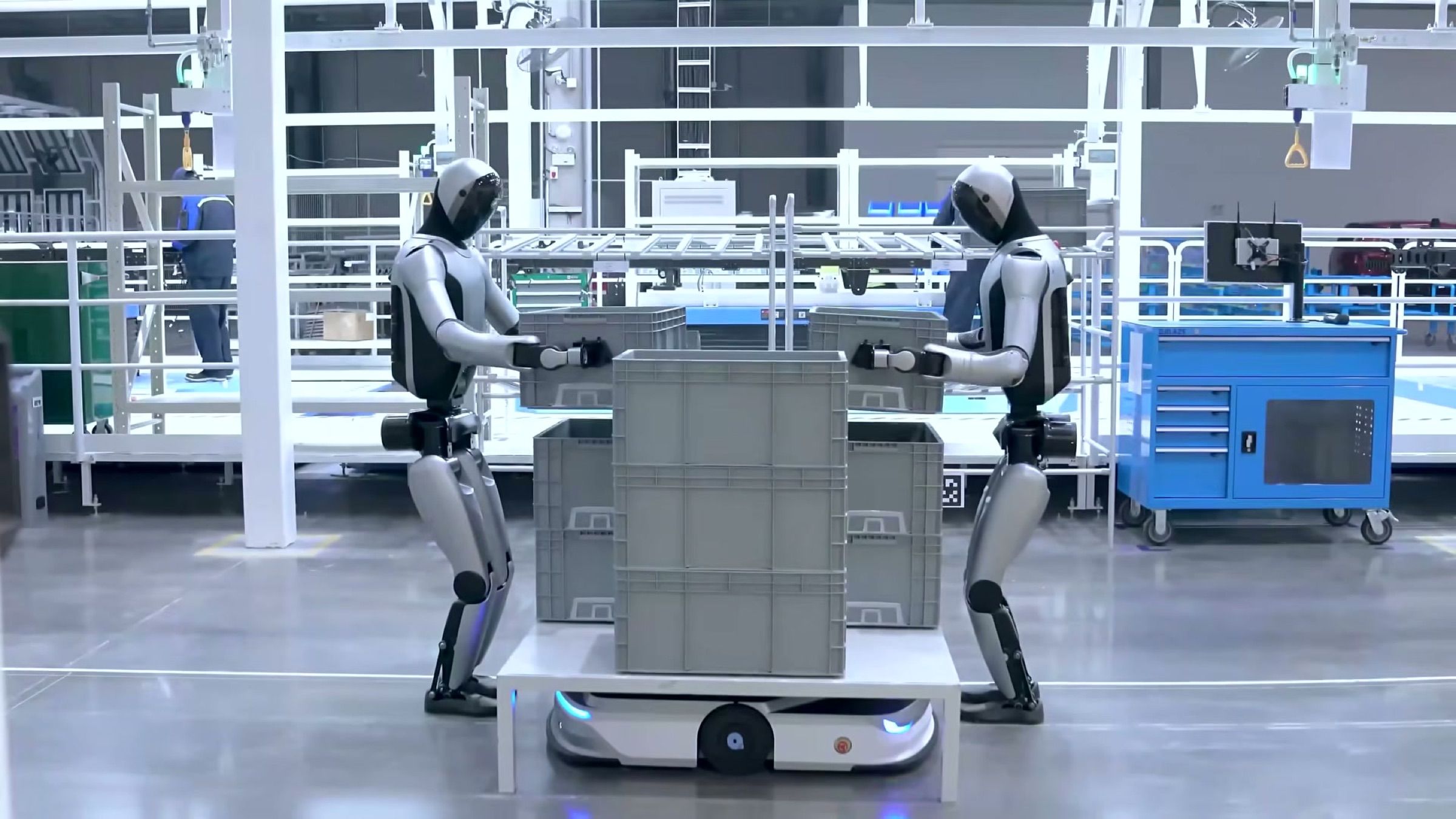 Two humanoid robots work together to load stacked totes onto an autonomous mobile robot on a factory floor.